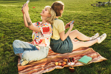 Image of smiling two women using mobile phones while resting on grass