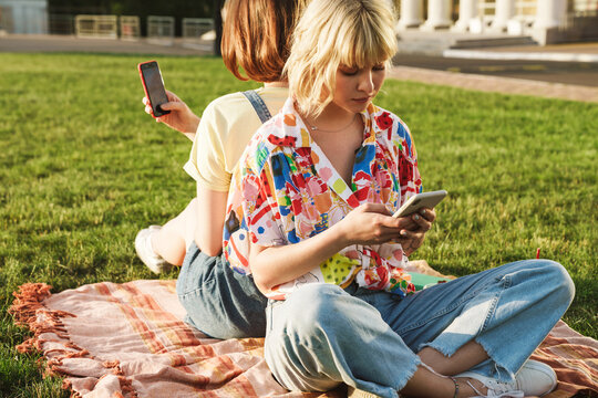Image of focused two women using mobile phones while resting on grass