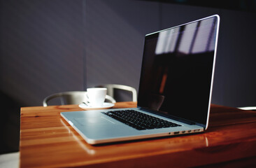 Portable net-book and cup of hot cappuccino lying on wooden table in contemporary coffee shop interior, freelance work via internet, open laptop computer and mug of cafe in modern office space