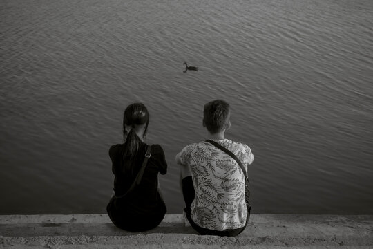 A view from the back of a young couple who is sitting on the steps in front of a pond and a duckling is swimming in front of them