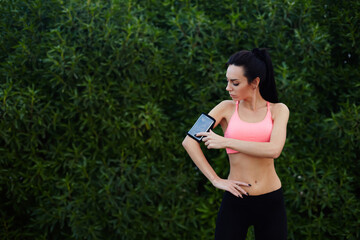 Naklejka na ściany i meble Sporty young woman standing in the park with big copy space for text while getting ready for workout,healthy fit runner setting her personal trainer application or stopwatch on cell phone before a run