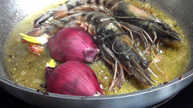 A zoom out shot of two tiger prawns and red onion cooking in melted garlic butter in a stainless-steel pan