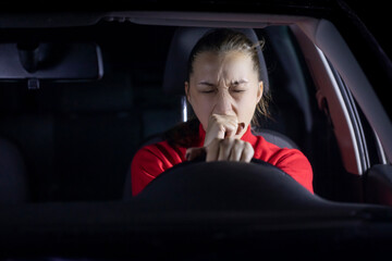 brunette long hair girl in red sweater driving car at night yawns tired holding steering wheel with...