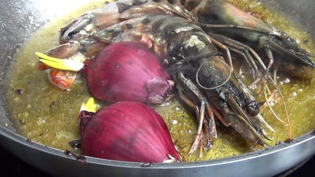 Caucasian white male hand puts one half of a red onion next to two tiger prawns which are cooking in melted garlic butter in a stainless-steel pan