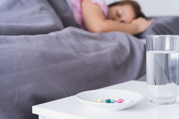 Fototapeta na wymiar Close up pill drugs in the dish and a glass of water on nightstand and female patient lying on the bed in the bedroom. Healthcare and medical pharmacy concept