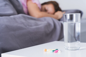 Fototapeta na wymiar Close up pill drugs and a glass of water on nightstand and female patient lying on the bed in the bedroom. Healthcare and medical pharmacy concept