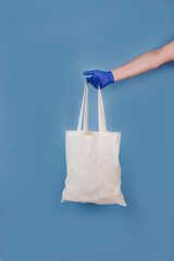 Vertical composition with man's hand in blue disposable protective gloves, holding white bag with food or goods donations on blue trendy background. Delivery or donation concept, mockup and copyspace