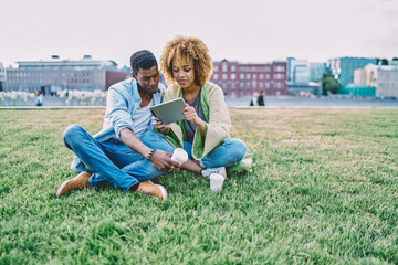 Happy couple in love spending time together in park on green grass and watching video online on modern touch pad device.Positive african american young man and woman doing shopping together on tablet