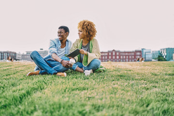 Positive african american hipster guy laughing while spending leisure time together with sceptical girlfriend with curly hair in park.Cheerful young people with touch pad sitting on green grass