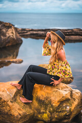 Caucasian blonde girl in a floral shirt, black shorts and straw hat in a natural landscape by the sea and rocks at sunset, lifestyle. Sitting on a rock in a natural lake