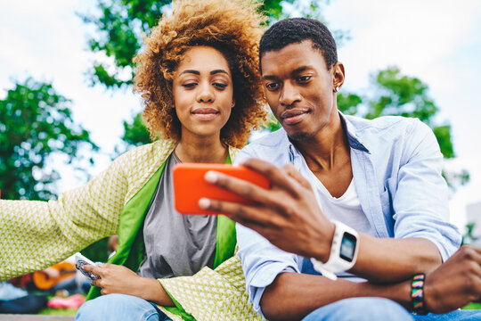 Two african american hipster friends dressed in casual wear watching video on website on modern smartphone using 4G internet connection.Couple in love making selfie photo on cellular resting outdoors