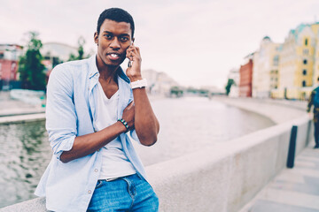 Portrait of positive afro american man dressed in stylish clothing having mobile conversation with friends on telephone connected to 4G.Handsome male looking at camera during talking on cellular