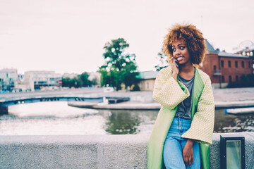 Young cheerful female person dressed in casual outfit contacting with friends in positive atmosphere using smartphone application.Cute happy hipster girl talking on cellphone while strolling outdoors