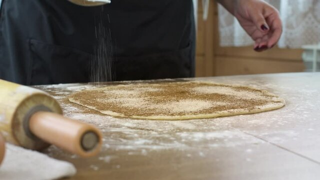 Close up female baker makes rolls from raw dough puff pastry for buns, woman sprinkles dough with sugar and cinnamon. Sweet pastries, bakery, dessert recipe, cooking at home, homemade concept