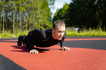A cute Caucasian guy in a black sports uniform stands in a plank position on a sports field in a Park on a summer day. Training of muscles and endurance.