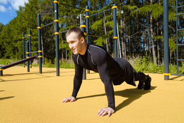 An athletic young man in a black elastic sports uniform stands in a plank position on the Playground in the Park. The concept of doing outdoor sports alone during a coronovirus epidemic.