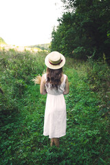 Girl in a hat and sundress outside the city. Young woman in a white summer board with a bouquet. Girl in a green summer field.