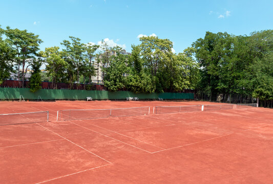 Empty Tennis Court On Sunny Summer Day. View From Above Of A Red Clay Tennis Court, Green Trees And A Blue Sky In The Background. Outdoor Sports Playground For Tennis. Copy Space For Text Or Design