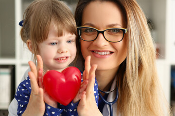 Happy mature female pediatrician hugging little kid during medical checkup in her office in the hospital. Doctor and child holding a toy heart