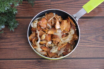 Fried chanterelles (roosters) with onions in a small green pan on a wooden table on a background of greenery, top view