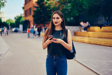 Charming brunette student strolling around city and booking online in website on modern telephone connected to 4G internet.Attractive pensive female blogger enjoying free time outdoors with gadget