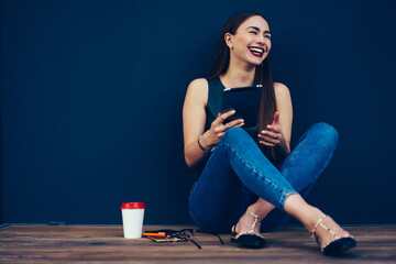 Cheerful female blogger dressed in trendy outfit having fun during online chatting with friends in social network.Emotional hipster girl loudly laughing while sitting next to wall with copy space
