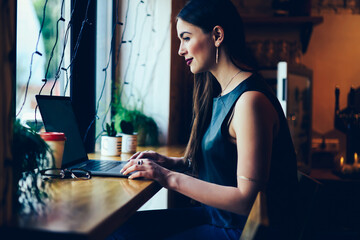 Charming positive skilled graphic designer sending email for customer while remotely working on laptop computer connected to internet.Beautiful female freelancer making remote job at coffee shop
