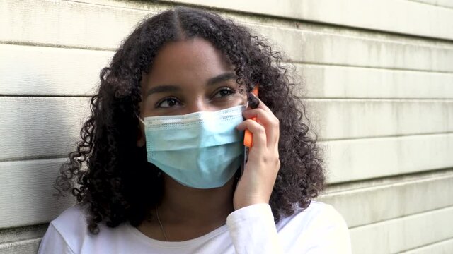Beautiful mixed race African American girl teenager young woman wearing a face mask during COVID-19 Coronavirus pandemic outdoors talking on her smartphone or cell phone