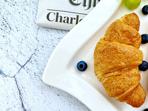 Closeup view of freshly baked croissants on a white plate with blueberries and grapes. Tasty breakfast with morning newspaper on a natural granite stone table. 