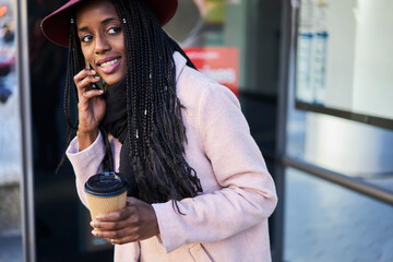 Cheerful young afro american female in trendy hat talking on phone standing outdoors waiting for taxi with coffee to go in morning getting to city downtown to meet with friends hanging out together