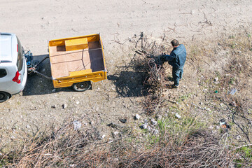 Fototapeta na wymiar The man cutting the dried braches gathered in a pile near the car with yellow cargo trailer on cart road. Top view.