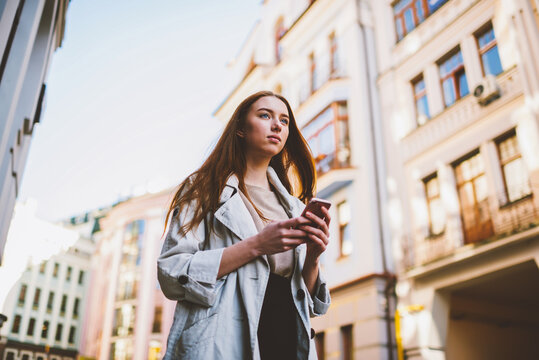 Gorgeous teenager dressed in trendy clothing looking away during walking at street. young hipster girl using application on smartphone connected to 4G and browsing interesting places to visit