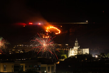 A fire caused by sparks from fireworks burns on the hillside above the city while firetrucks line the ridge above and people set off firecrackers below.