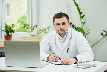 A caucasian doctor in a white lab coat is doing paperwork near a laptop in a hospital. A therapist is waiting for a patient in a doctor's office.