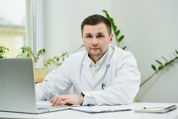 A serious caucasian doctor in a white lab coat was distracted while searching medical information on a laptop in a hospital. A therapist near a computer in a doctor's office.