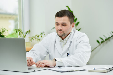 A serious caucasian doctor in a white lab coat is entering the patient’s medical history into a...