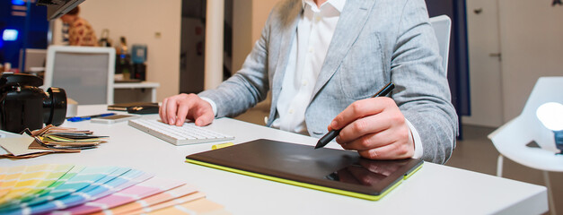 Photographer-designer in a gray jacket works at a table, holds a stylus of a graphic tablet in the...