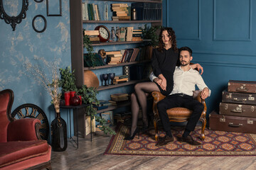 Obraz premium Horizontal full length portrait of young elegant couple in old antique room. Man and woman posing near bookshelf with a lot of ancient antiques. Dark curly woman sitting on the armrest with husband.