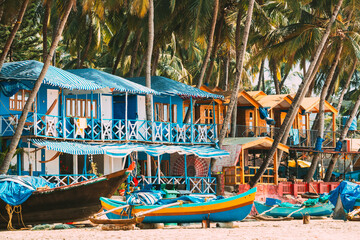 Fototapeta na wymiar Canacona, Goa, India. Fishing Boat And Famous Painted Guest Houses On Palolem Beach Against Background Of Tall Palm Trees In Sunny Day