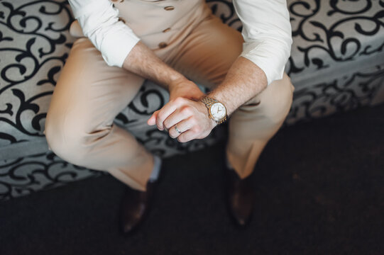 The groom in a beige suit and waistcoat sits on the couch and straightens the clock. Concept of waiting bride. Wedding details.