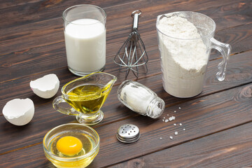 Measuring cup with flour, glass of milk, broken egg and salt, metal whisk on table.