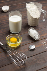 Measuring cup with flour, glass of milk, broken egg and salt, metal whisk on table.