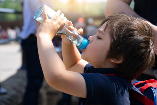 Cute Little Boy Drinking Colding From Clear Glass Bottle In The Hot Sunny Day On Summer, Kid Boy Drink Soda Or Soft Drink Sitting In Street With Crowd Of People Around