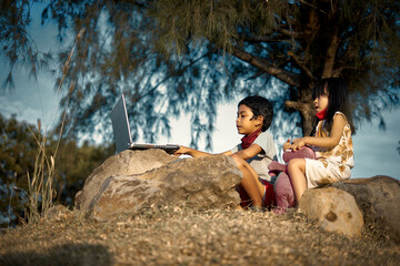 A boy and his little cute sister sitting on a rock and study using laptops under the tree.  New normal for Education concept