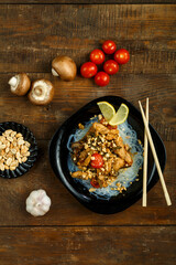On a wooden table is a plate with chicken and vegetables in pad Thai sauce and funchose