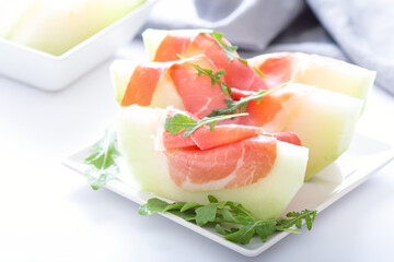 Traditional Italian or Spanish appetizer fresh melon with prosciutto or jamon and green herbs on white table, top view, light mediterranean cuisine dish