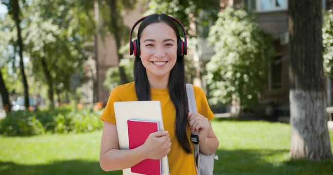Portrait of cute asian student girl with earphones, backpack, red notebook and gray laptop looking at camera and smiling with green park at background.