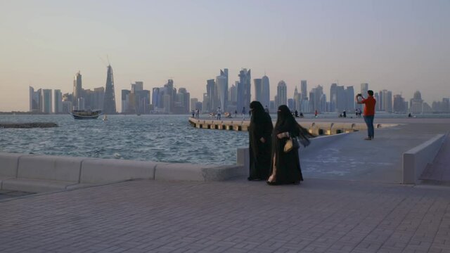 Doha corniche sunset shot showing people walking on Cor niche promenade with Doha skyline and Dhow in the Arabic gulf in the background
