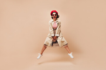 Emotional woman in beret and coat jumping on beige background. Bright girl with dark hair in sunglasses and white sneakers posing..