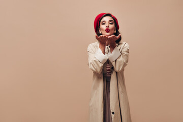 Charming woman in red beret and trench blows kiss on beige background. Stylish lady with dark hair in long light coat posing on camera..
